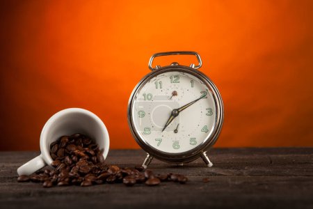 Photo for Coffee beans scattered on a surface near a small cup and an alarm clock on a table, isolated on orange background - Royalty Free Image