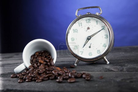 Téléchargez les photos : Grains de café dispersés sur une surface près d'une petite tasse et un réveil sur une table, isolé sur fond bleu - en image libre de droit