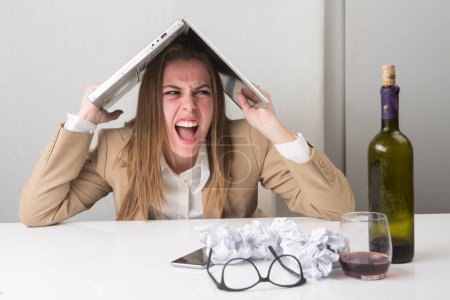 Photo for Manager woman in jacket sitting in her workplace is desperate and carries the laptop on her head, with a bottle of wine next to her - Royalty Free Image