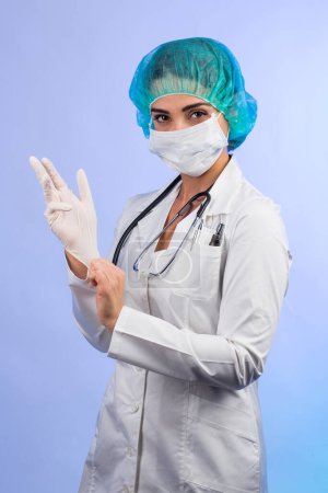 Foto de Doctora en bata blanca, mascarilla blanca, gorro verde para el cabello tiene la intención de ponerse guantes de látex, aislados sobre fondo blanco - Imagen libre de derechos