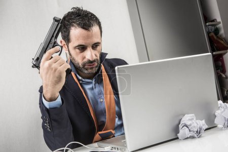 Photo for Dark-haired manager dressed in a suit and upset and sitting in his office and looks with hatred at his computer holding a gun in his hand - Royalty Free Image