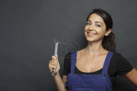 Photo for Beautiful girl with worker work tools in hand and blue overalls gurada curiously, isolated on black background - Royalty Free Image