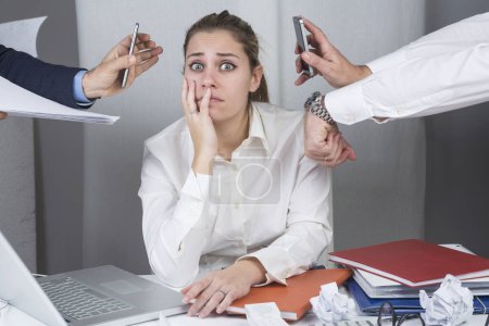 Photo for Manager woman sitting in her desk is surrounded by hands and commitments and looks depressed - Royalty Free Image