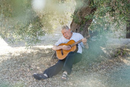 Téléchargez les photos : Homme de quatre-vingts ans mélancolique jouant une mélodie assise à l'ombre d'un vieil olivier à la campagne - en image libre de droit