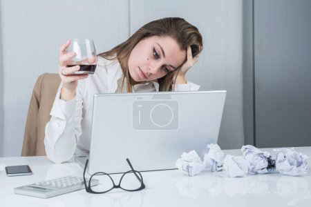 Foto de Chica rubia con chaqueta está sentada en su escritorio de la oficina delante de su computadora portátil de trabajo, mientras que ella se distrae con el vino tinto - Imagen libre de derechos
