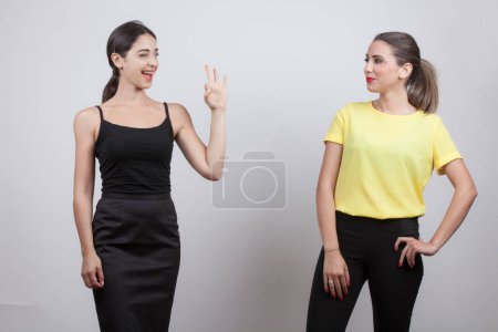 Foto de Dos chicas, de las cuales una morena vestida de negro y la otra rubia vestida de faja amarilla intercambian un signo de comprensión y risa, aislada sobre fondo gris - Imagen libre de derechos