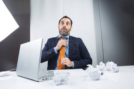 Photo for Manager in elegant suit sitting in his workplace in front of his laptop arranges the orange carvatta with funny face - Royalty Free Image