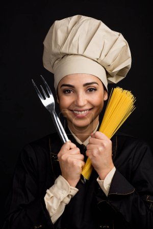 Téléchargez les photos : Un cuisinier souriant avec un chapeau de chef tient des spaghettis crus et une fourchette sculptée, isolés sur fond noir - en image libre de droit