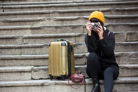 Téléchargez les photos : Jeune touriste en vêtements d'hiver assis sur les marches avec le chariot à proximité, prend une photo - en image libre de droit