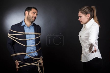 Photo for Man in jacket tied with a rope and desperate in front of a blonde girl who looks at him angry. the couple argues furiously isolated on black background. - Royalty Free Image