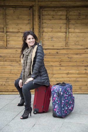 Photo for Tourist in winter clothes is sitting on top of the suitcases in front of a wooden kiosk in a square - Royalty Free Image