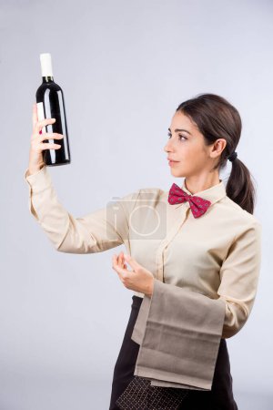 Photo for Beautiful and elegant waitress with shirt and red bow tie and napkin in her arm and ready to serve wine from a bottle, isolated on a light background - Royalty Free Image