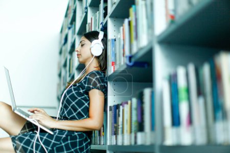 Téléchargez les photos : Jeune fille brune avec casque fonctionne sur l'ordinateur assis parmi les étagères dans une bibliothèque - en image libre de droit