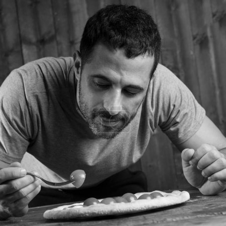 Foto de Hombre de pelo oscuro con barba está a punto de comer una pizza con tomates cherry, aislado sobre fondo de madera, blanco y negro - Imagen libre de derechos
