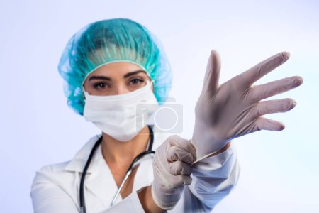 Photo for Female doctor in white coat, white face mask, green bonnet for hair is intent on putting on latex gloves, isolated on white background - Royalty Free Image