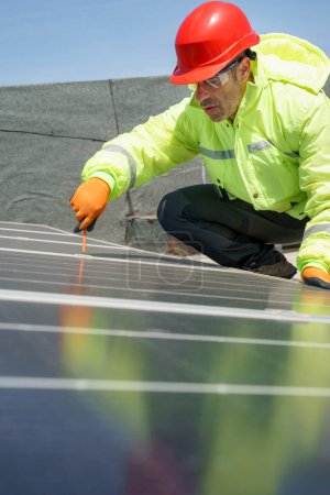 Foto de Trabajador con casco protector rojo, mono especialista y gafas técnicas trabaja en un sistema fotovoltaico instalado en el techo de un edificio - Imagen libre de derechos