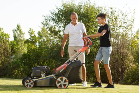 Téléchargez les photos : Père avec fils tondre l'herbe avec la tondeuse dans le jardin de la maison - en image libre de droit