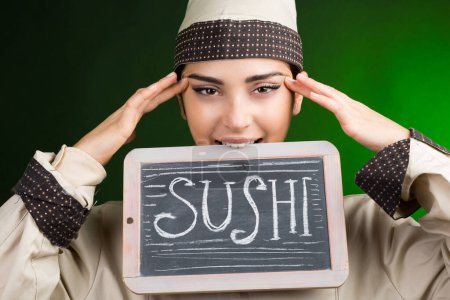Foto de Cocinero europeo en uniforme sostiene una pizarra con sushi escrito en él, aislado sobre un fondo verde. - Imagen libre de derechos