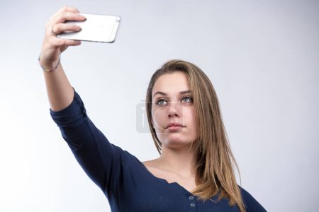 Photo for Blonde girl in blue shirt takes a selfie with a smartphone , isolated on white background - Royalty Free Image