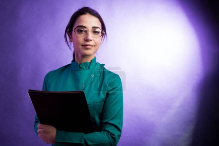 Foto de Doctora en bata de laboratorio verde sosteniendo una carpeta negra y gafas mientras mira severamente, aislada sobre fondo púrpura - Imagen libre de derechos