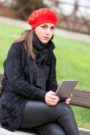 Foto de Chica vestida de negro con un auricular rojo sentado en un banco en un parque - Imagen libre de derechos
