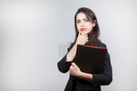 Photo for White girl with dark hair dressed in black jacket holds a black folder in hand and looks puzzled isolated on white background - Royalty Free Image