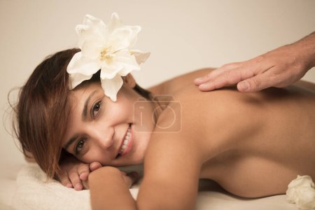 Photo for Beautiful woman with bob hair and a lotus flower on her head relaxes on a spa bed lying on her stomach ready to receive a back massage - Royalty Free Image