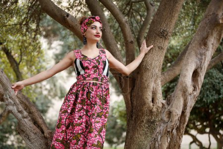 Photo for Beautiful smiling girl with red lipstick dressed in a colorful dress and a headband of the same material, stands in the middle of a natural park. - Royalty Free Image