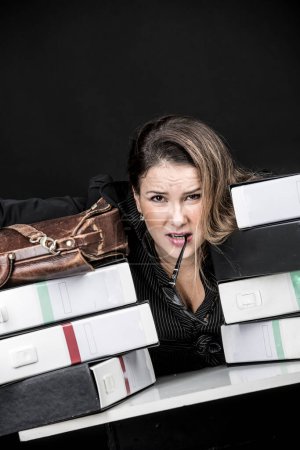Photo for Blonde secretary with eyeglasses, dressed in black and desperate and worried amidst all the messy documents in her office, isolated on black background - Royalty Free Image
