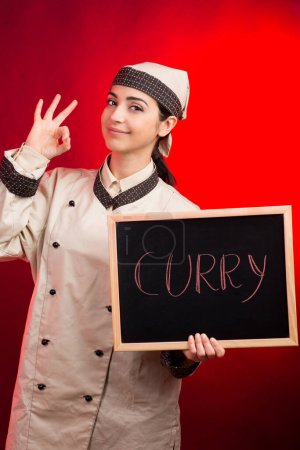 Photo for Cook makes the ok sign with holding a blackboard with "Curry" written on it, isolated on red background - Royalty Free Image