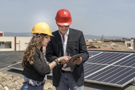 Foto de Pareja de ingenieros con casco protector comprueban el estado de un sistema fotovoltaico en el techo de una casa utilizando una tableta de ingenieros sistema fotovoltaico - Imagen libre de derechos