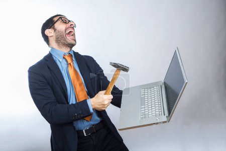 Téléchargez les photos : Homme aux cheveux bruns avec barbe, vêtu d'un costume chemise, veste et cravate orange, prend un ordinateur portable dans sa main avec un marteau, isolé sur fond blanc - en image libre de droit