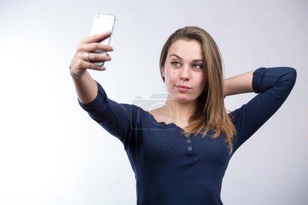 Téléchargez les photos : Blonde fille en chemise bleue prend un selfie avec un smartphone, isolé sur fond blanc - en image libre de droit