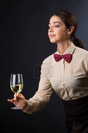 Téléchargez les photos : Belle et élégante serveuse avec chemise et nœud papillon rouge est prête à offrir un verre de vin à un client, isolé sur un fond noir - en image libre de droit