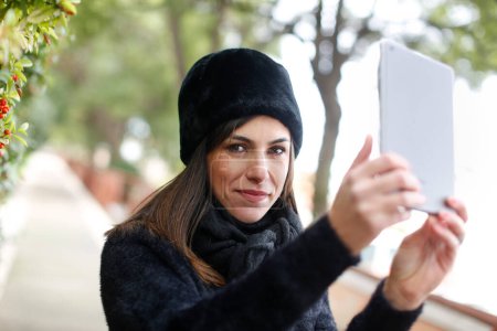 Foto de Chica en un sombrero de piel negro toma una selfie con una tableta en un parque - Imagen libre de derechos