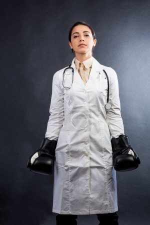 Téléchargez les photos : Femme médecin en manteau blanc et gants de boxe A l'expression de quelqu'un qui ne veut pas abandonner, isolé sur fond gris - en image libre de droit