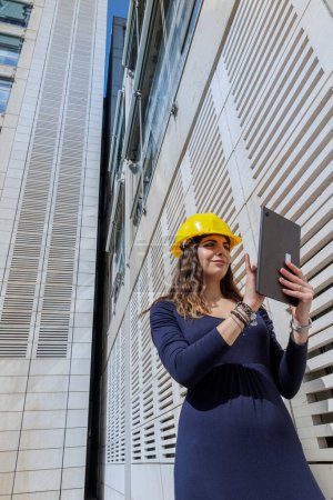 Foto de Mujer elegantemente vestida con casco protector amarillo, sosteniendo su tablet en la mano en un contexto urbano moderno - Imagen libre de derechos