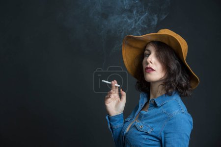 Photo for Brunette girl with denim shirt and cowboy hat, smokes a cigarette producing a lot of smoke, isolated on gray background - Royalty Free Image