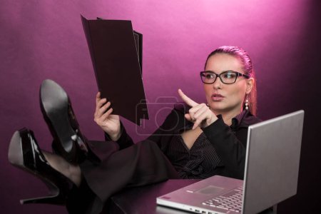 Téléchargez les photos : Femme manager avec les cheveux collectés portant des lunettes, habillé en costume d'affaires noir travaillant sur ordinateur avec les pieds sur la table, isolé sur fond rouge - en image libre de droit
