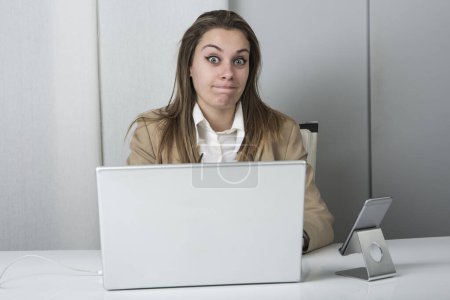Téléchargez les photos : Gestionnaire blonde habillé occasionnel assis dans son lieu de travail devant son ordinateur portable et son smartphone fait un drôle de visage - en image libre de droit