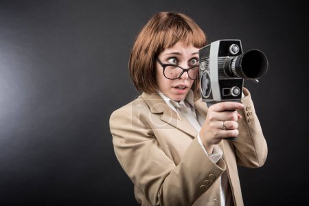 Photo for Girl with glasses, bob hair and beige jacket, uses an old movie camera looking at her with funny expression of uncertainty, isolated on black background - Royalty Free Image