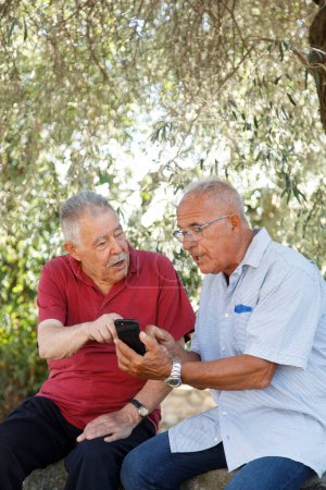 Photo for Couple of Elderly friends look at a cellphone sitting in an outdoor park - Royalty Free Image