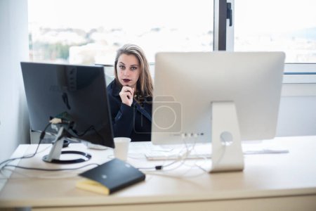 Photo for Blonde office worker sitting on the working chair near her desk looks with serious expression - Royalty Free Image