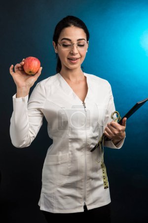 Foto de El dietista de capa blanca sostiene en la mano un medidor para medir el cuerpo humano y una manzana que muestra orgullosamente, aislada sobre fondo verde - Imagen libre de derechos