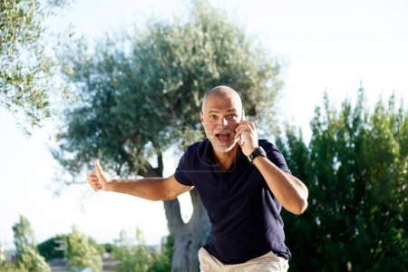 Foto de Un hombre de 50 años con una camisa negra y pantalones cortos de colores claros está hablando animadamente por teléfono en su patio trasero. - Imagen libre de derechos