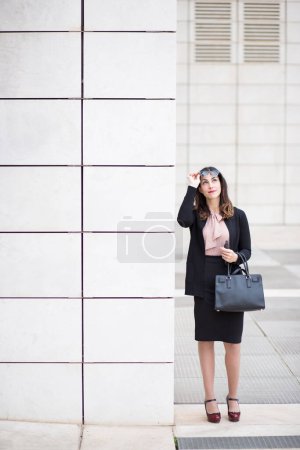 Téléchargez les photos : Brunette gestionnaire, vêtue d'un costume d'affaires noir posant dans une rue - en image libre de droit