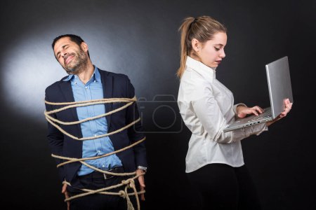 Foto de Hombre con chaqueta atada con una cuerda está desesperado delante de una chica rubia que controla el ordenador portátil, aislado sobre fondo negro. - Imagen libre de derechos