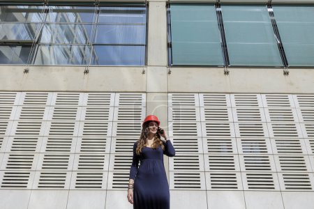 Foto de Mujer elegantemente vestida de rojo sombrero duro habla con expresión seria en el teléfono en el entorno urbano moderno - Imagen libre de derechos