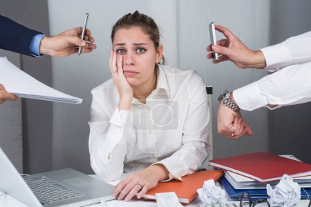 Photo for Manager woman sitting in her desk is surrounded by hands and commitments and looks depressed - Royalty Free Image
