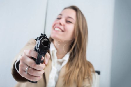 Photo for Blonde girl dressed in a beige jacket is sitting at her workstation she is holding a gun in her hand ready to use it - Royalty Free Image